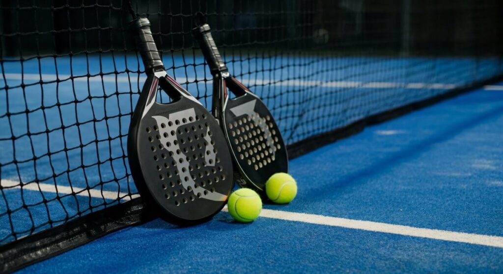 Two padel rackets and yellow balls beside a net on a blue court